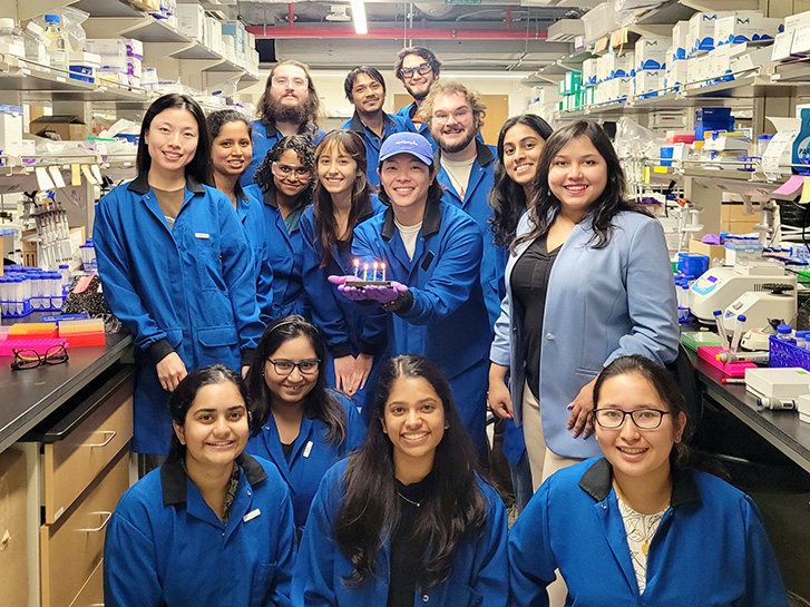 A group of people in blue uniforms pose for a photo inside a lab; a person in the center of the group holds a flat item with four lit candles